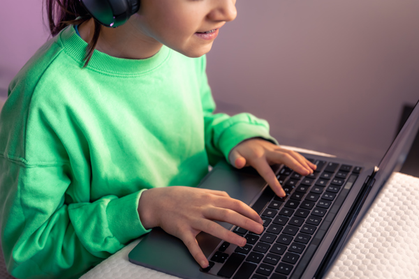Young person in a green sweatshirt wearing headphones, typing on a laptop.