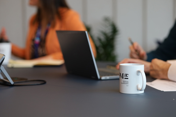 Close-up of a coffee mug on a table with an open laptop and papers, two people in the background writing and holding a pen.